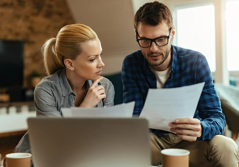 Hombre y mujer sentados frente a una computadora portátil revisando papeles y analizando finanzas domésticas en un ambiente hogareño.